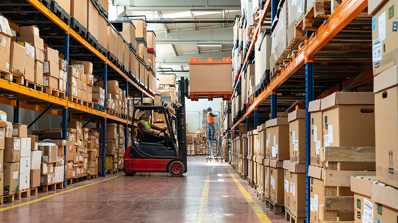 Warehouse worker driver in uniform loading cardboxes by forklift stacker loader